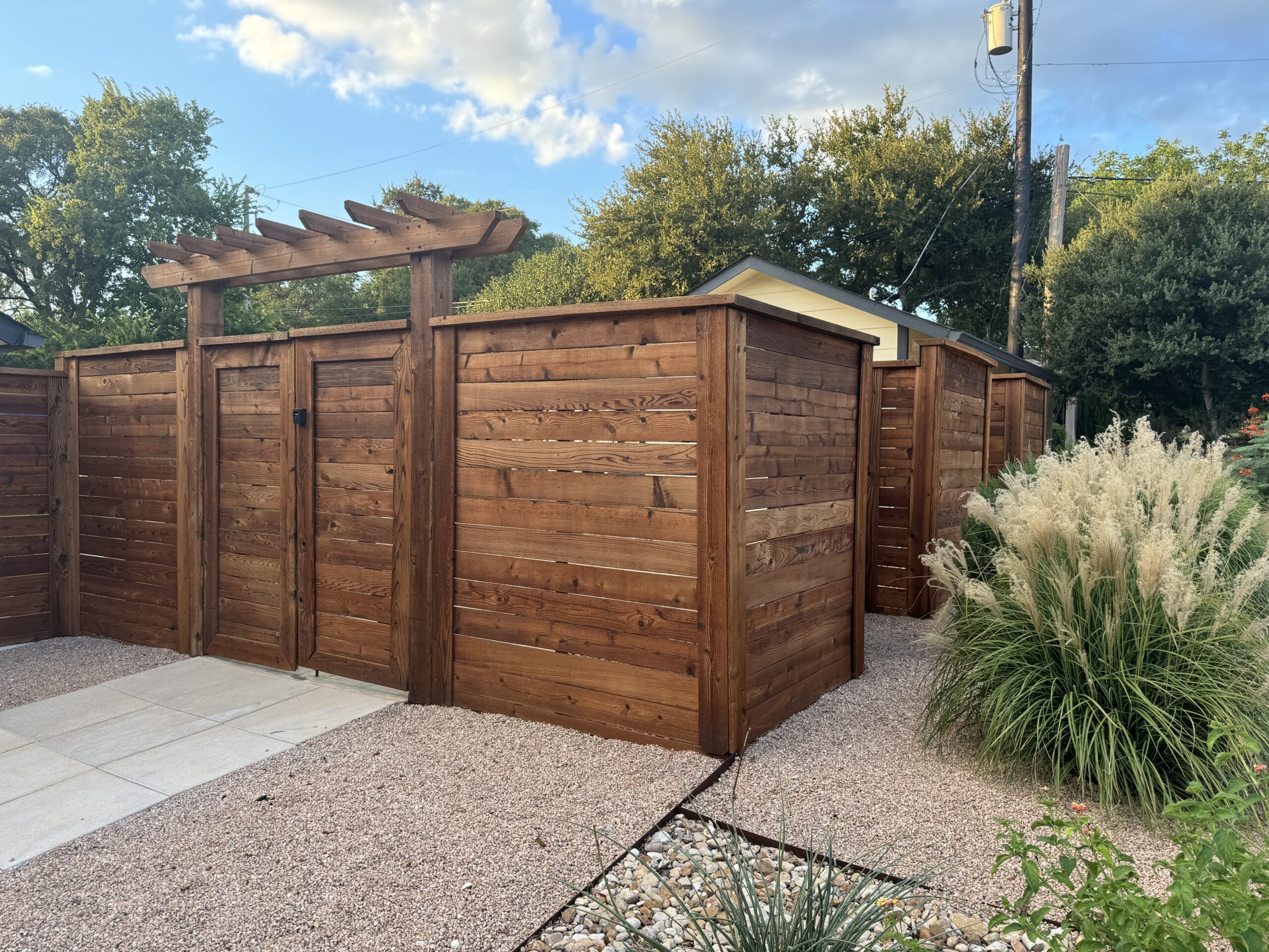 Wooden fence with a gate on a gravel path, surrounded by lush greenery and under a partly cloudy sky.
