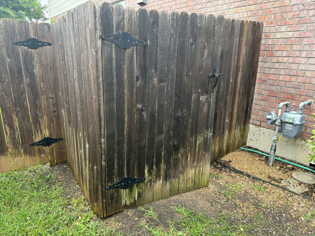Wooden fence corner with gate near a brick wall and a gas meter. The fence has metal hinges and shows signs of weathering, with green moss at the bottom. Grass grows at the base.