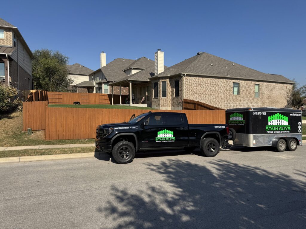 A black pickup truck and trailer with "Stain Guys" branding are parked on the street in front of a house with a newly stained wooden fence.