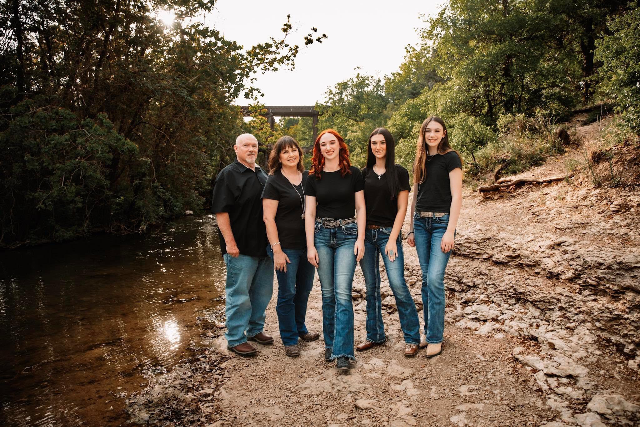Five people pose together on a rocky path beside a creek, surrounded by trees, all wearing black tops and blue jeans.