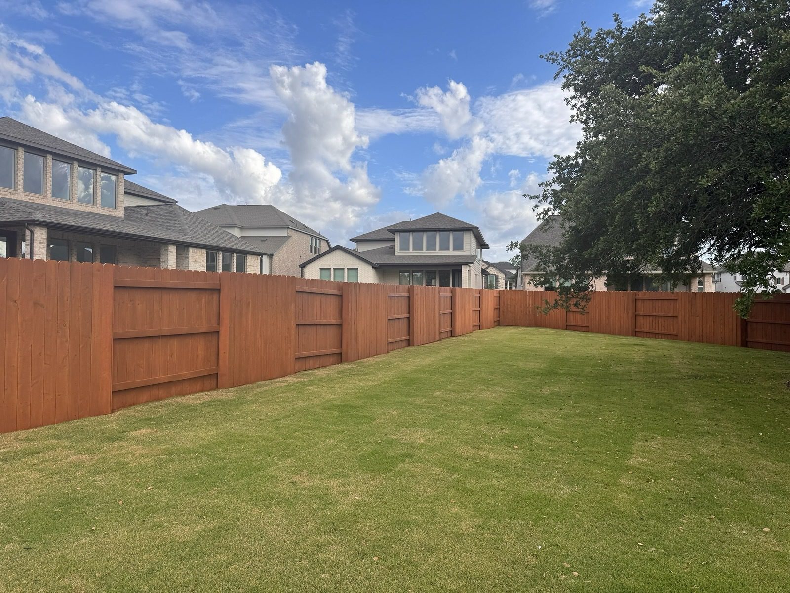 MEd18058fc40f5db7637f2ed5e7da24c97 A backyard with a well-kept green lawn, bordered by a tall wooden fence, with several modern houses and a large tree visible in the background under a partly cloudy sky.