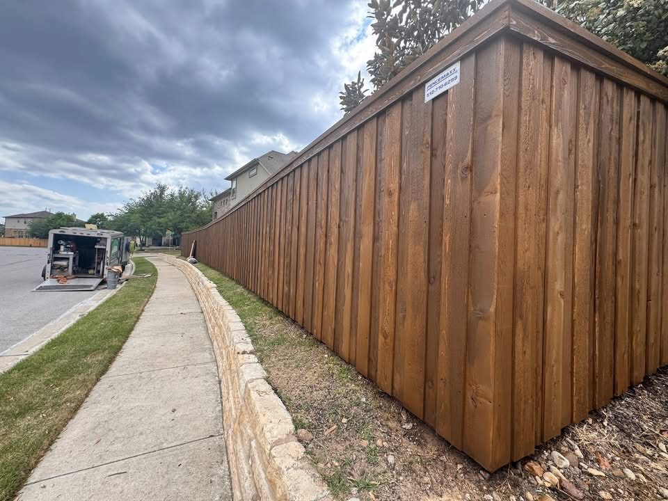 coffee-brown A tall wooden privacy fence runs along a sidewalk next to a suburban street under a cloudy sky, with a trailer parked in the background.