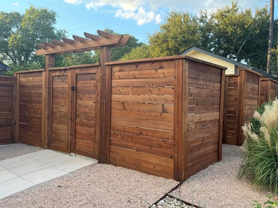 dark-walnut Tall wooden privacy fence with a gate and pergola overhang, surrounding a yard area with gravel and ornamental grass in the foreground.