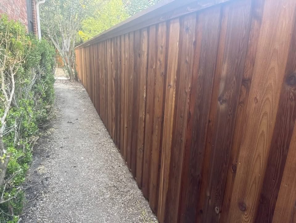 leatherwood A newly built wooden fence runs alongside a narrow dirt path bordered by green shrubs and a red brick building.