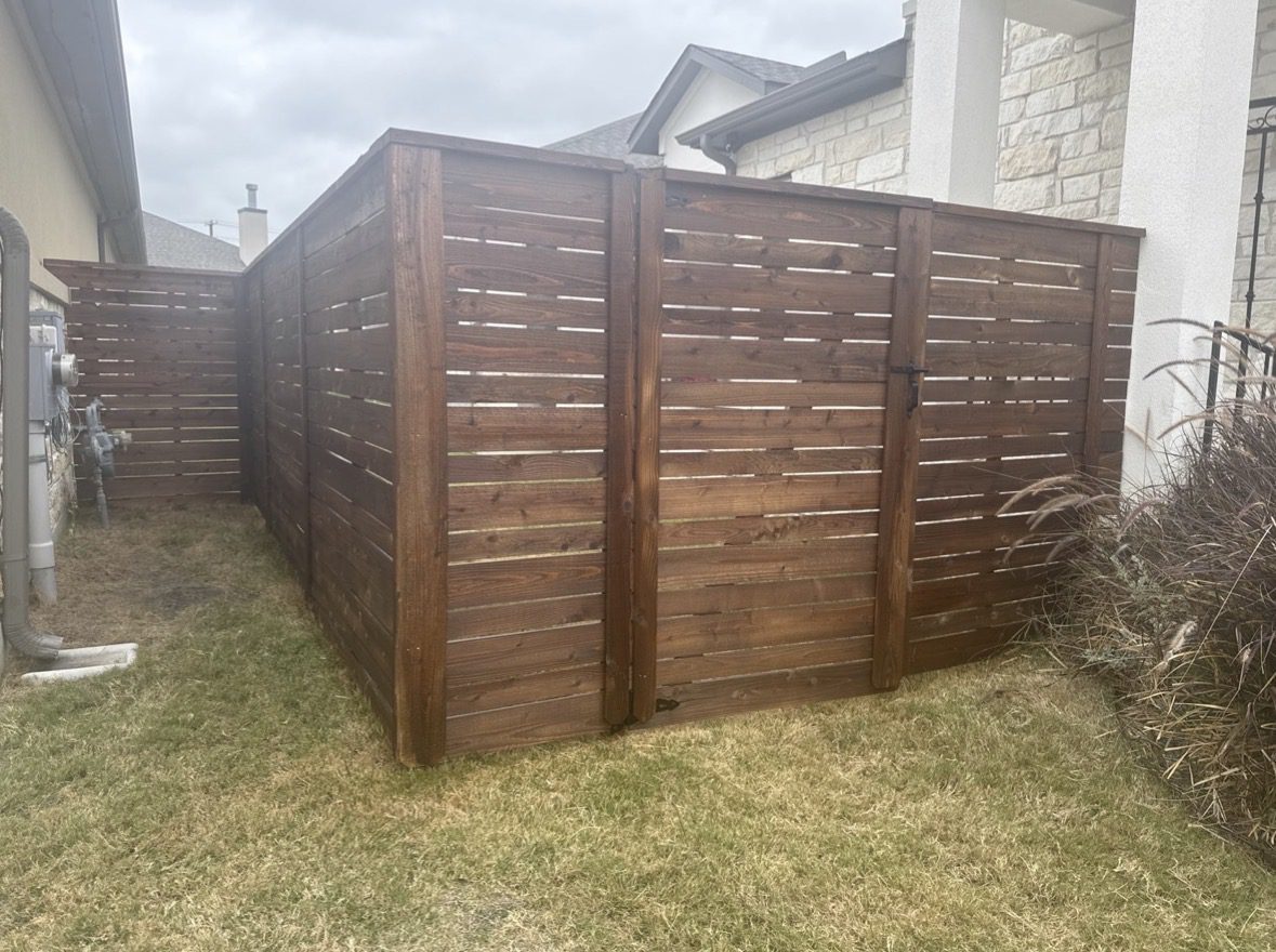 Screenshot A wooden privacy fence encloses a section of a grassy yard between two houses, with a gate visible in the front panel.