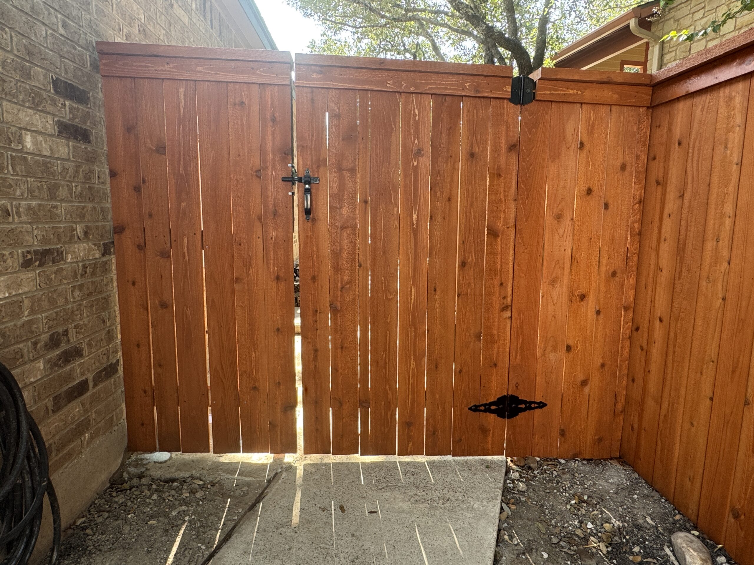 sierra A wooden fence gate with a black metal latch and hinge is closed between a brick house and another wooden fence. Sunlight casts shadows on the concrete below.