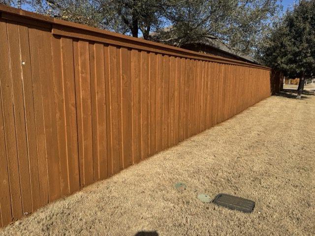 slide1 A long, wooden privacy fence runs along the edge of a dry, yellowish lawn with a few utility covers and trees in the background.