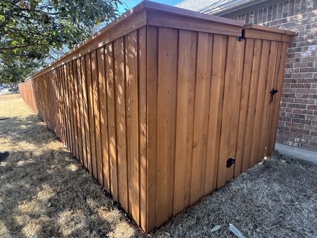 slide2 A long wooden privacy fence with a gate stands next to a brick house on a dry, patchy lawn under a tree.