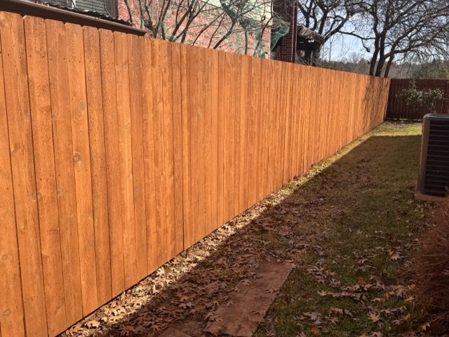 slide3 A tall, wooden privacy fence runs alongside a grassy yard with fallen leaves, bordered by houses and trees in the background.