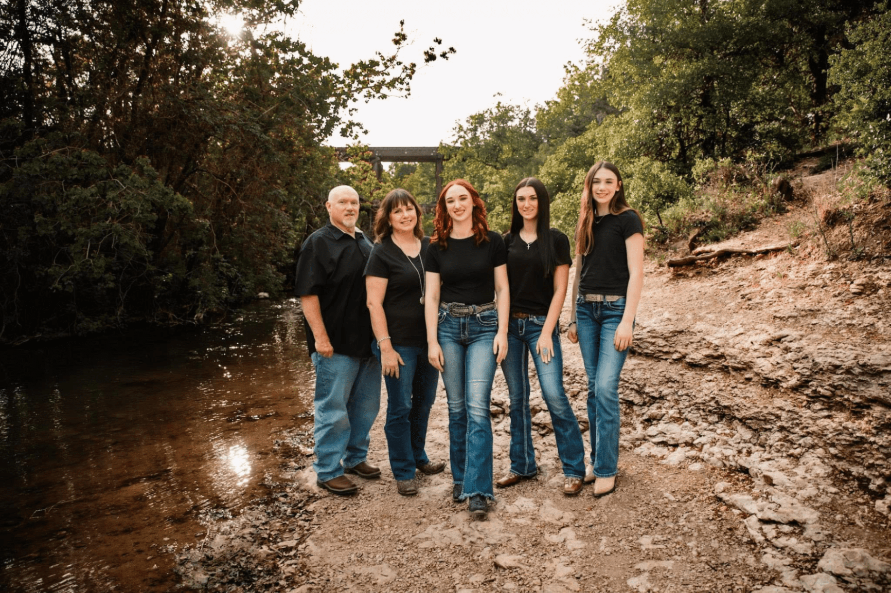 team Five people stand by a rocky creekside, all wearing black tops and blue jeans, with trees and a small bridge in the background.