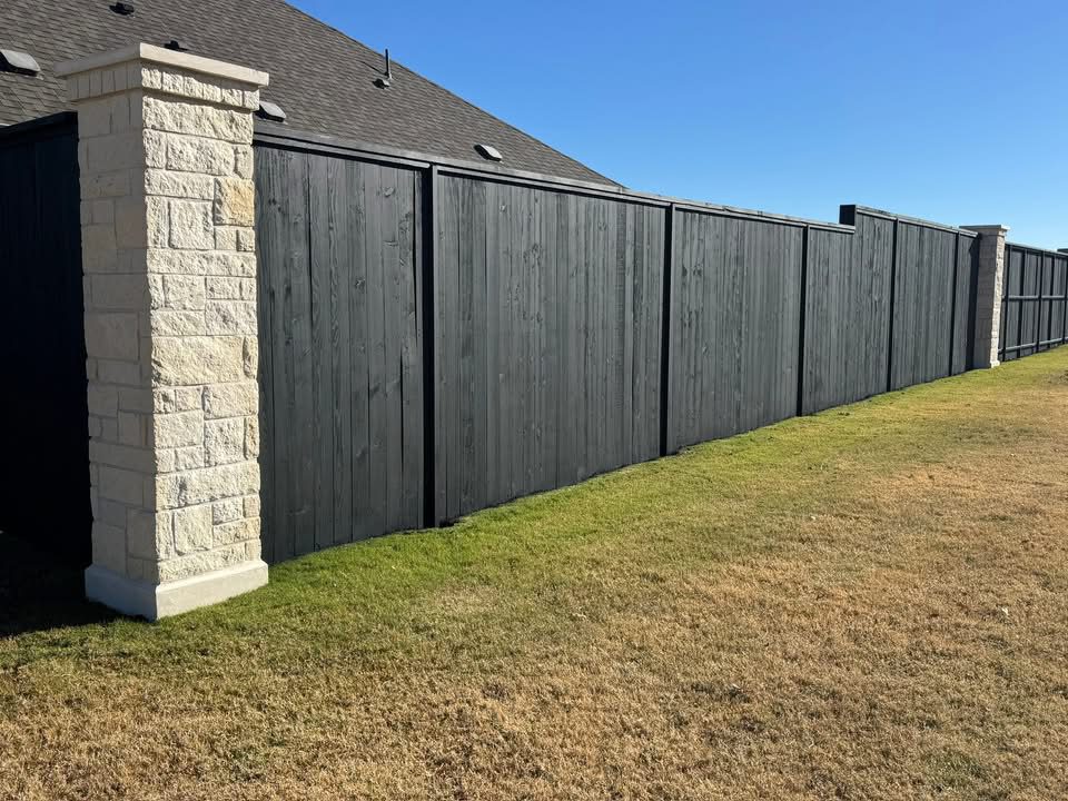 water-based-2 A tall wooden privacy fence with stone pillars stands alongside a house, bordered by a grassy yard and dry, brown grass under a clear blue sky.