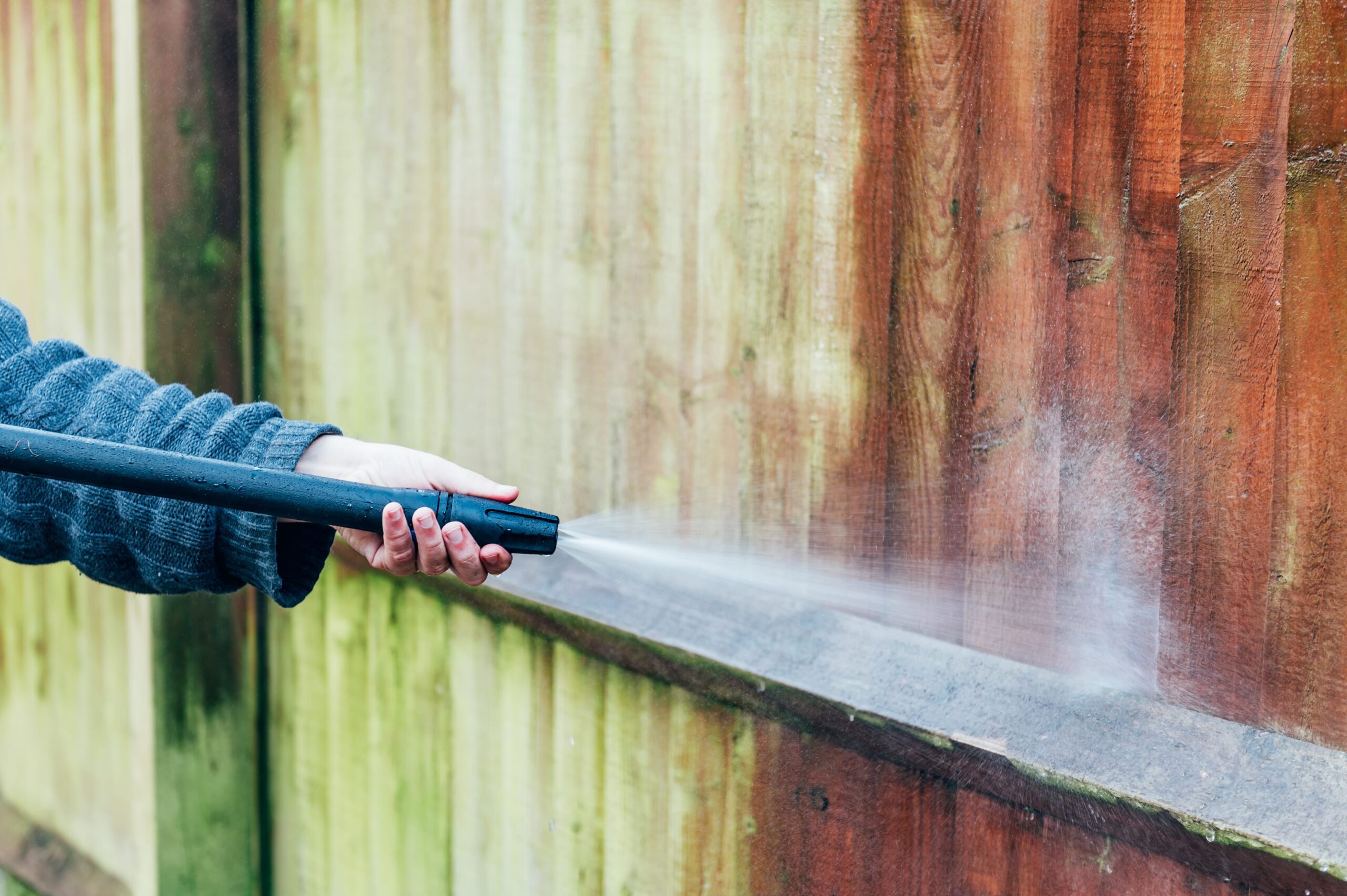 A person uses a pressure washer to clean a wooden fence, removing green moss and dirt to reveal the original wood color.
