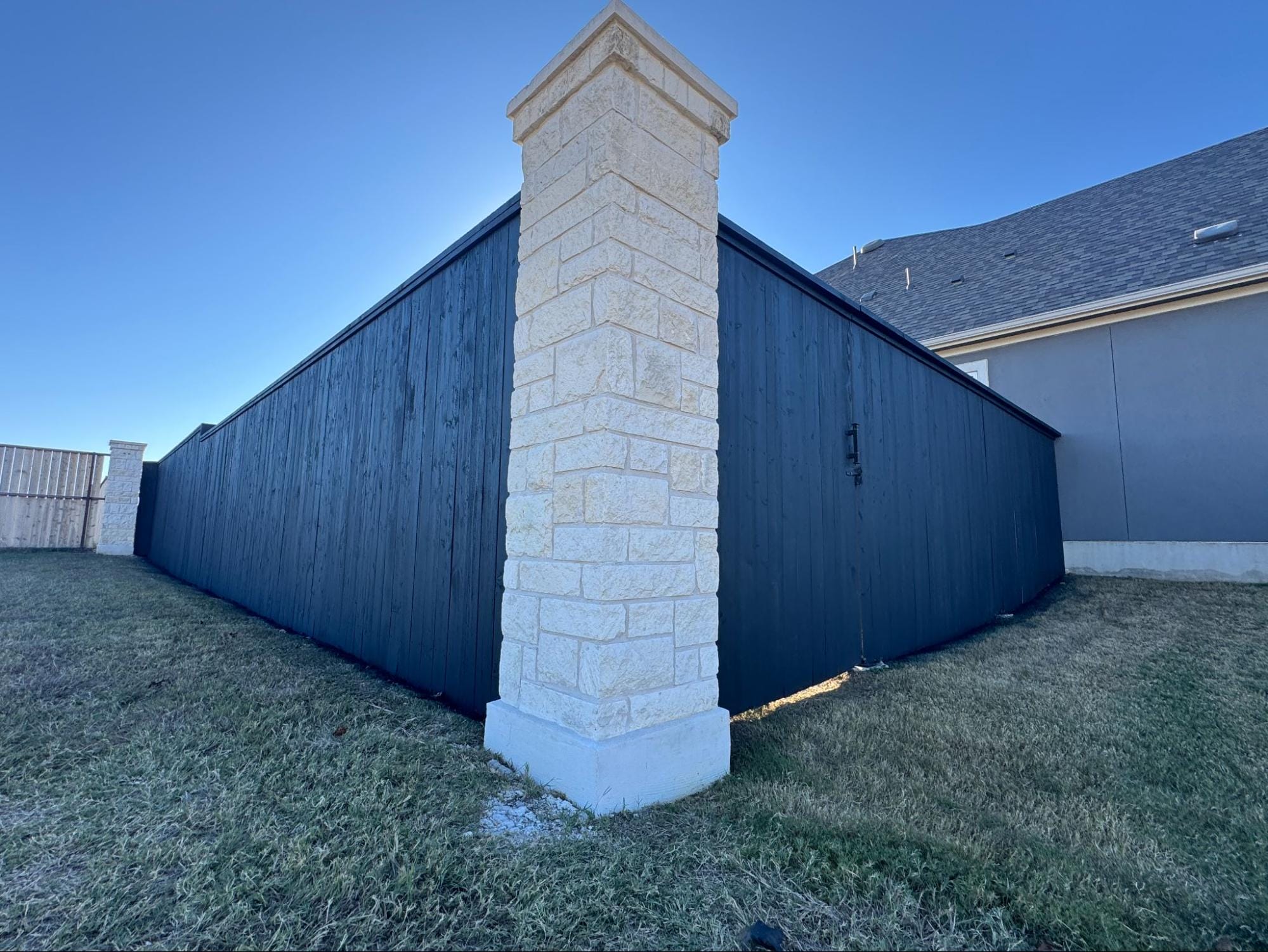 A stone pillar stands at the corner of a black wooden fence in a grassy yard next to a gray house under a clear blue sky. A stone pillar stands at the corner of a black wooden fence in a grassy yard next to a gray house under a clear blue sky.