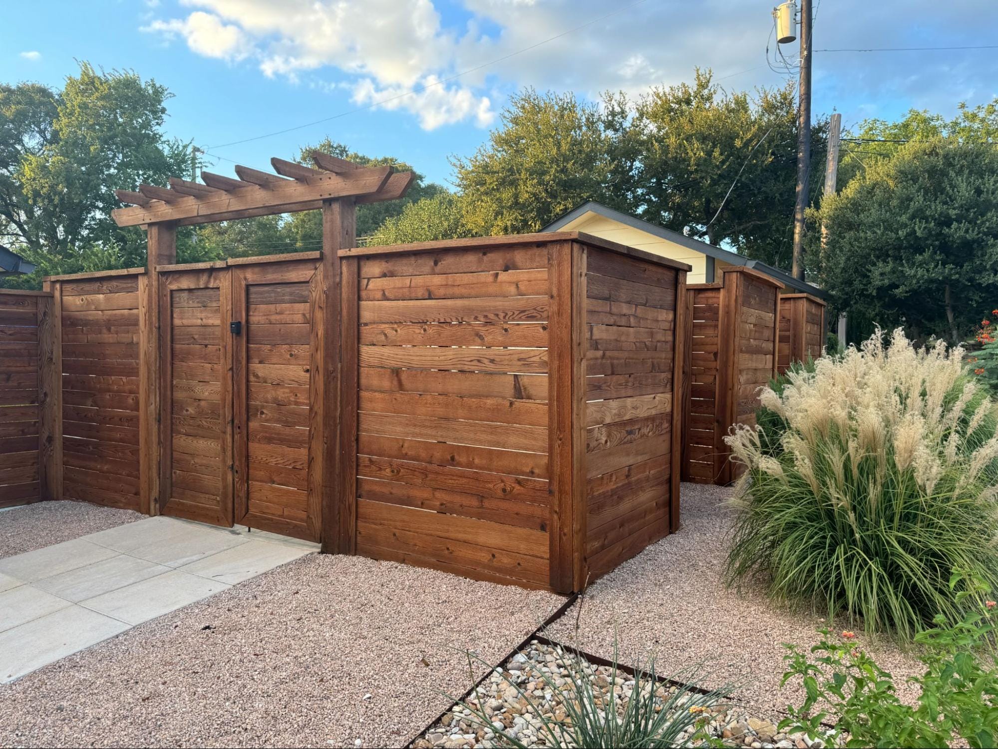 A wooden privacy fence with a pergola structure above the gate stands beside ornamental grasses and gravel landscaping under a partly cloudy sky.