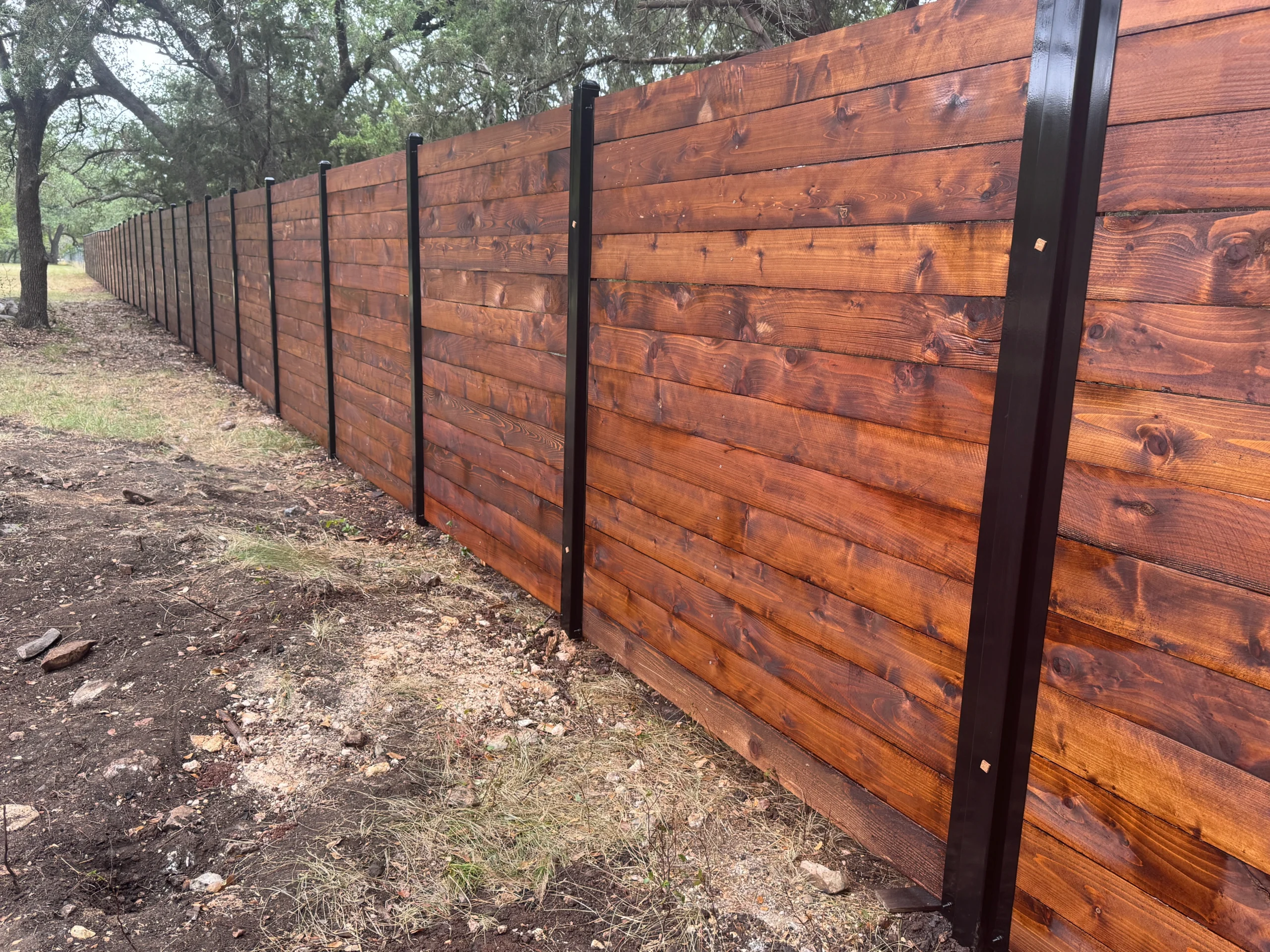 A long wooden fence with vertical black metal posts stands on a grassy area with trees in the background, expertly maintained by a local fence staining contractor.