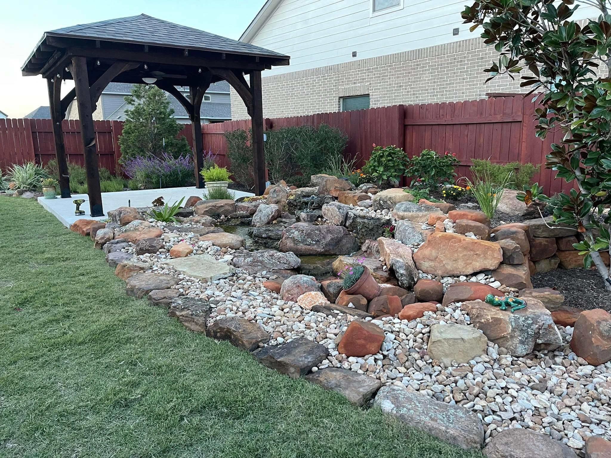 A landscaped backyard with a wooden gazebo, a rock garden with various-sized stones, small plants, and a red wooden fence in the background showcases the expert touch of a fence staining contractor.