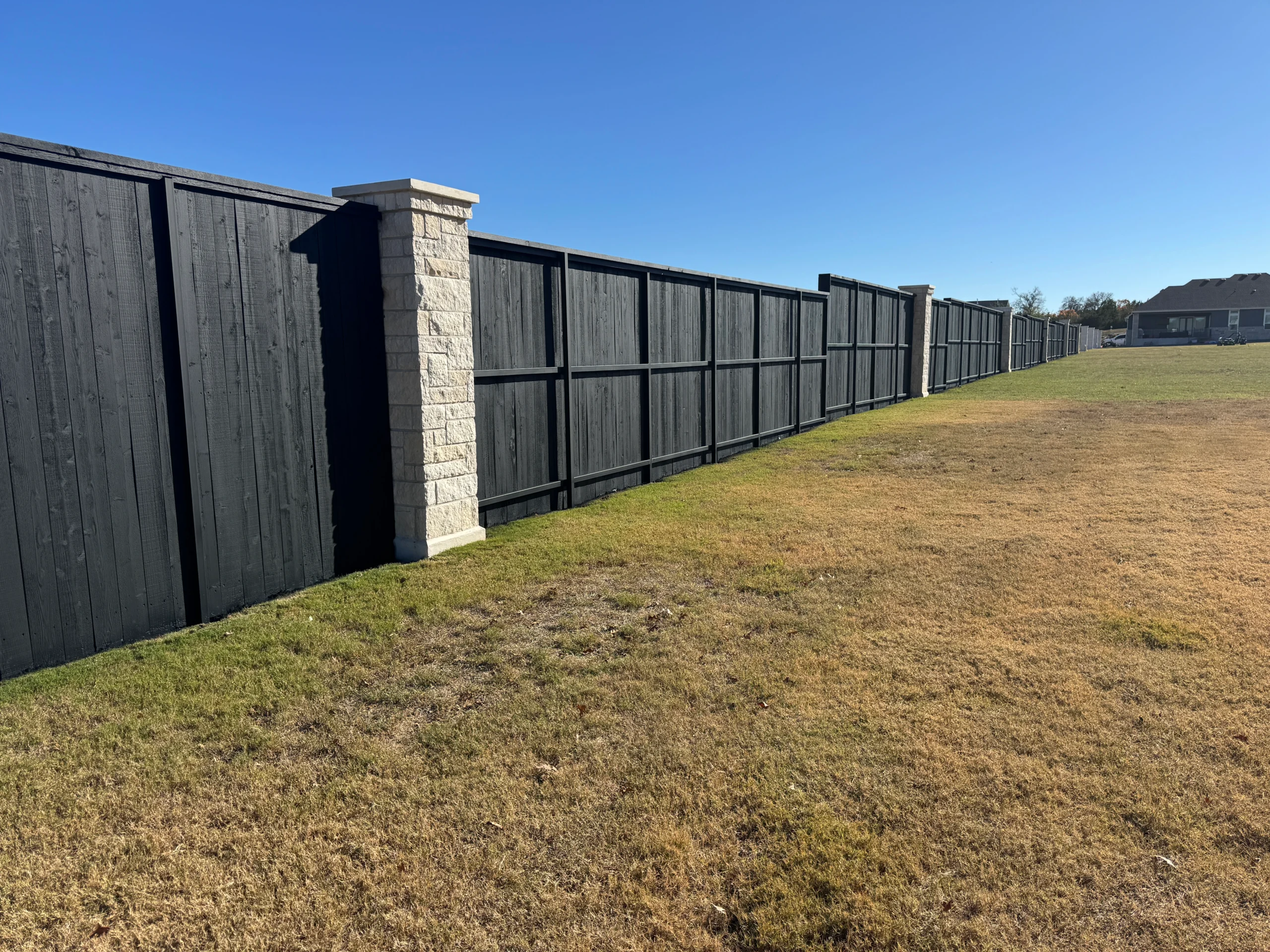 A long black wooden fence, expertly finished by a skilled fence staining contractor, with stone pillars runs along the edge of a grassy field under a clear blue sky.