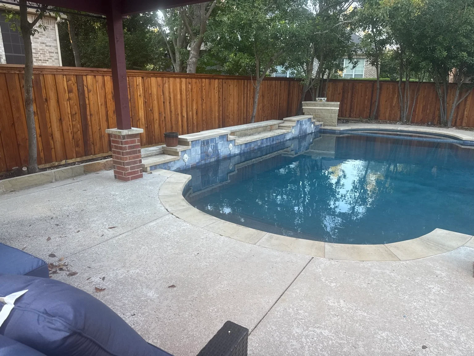 Backyard swimming pool with stone edge, tiled steps, wooden fence, and patio area with outdoor seating. Trees line the perimeter outside the fence, highlighting the craftsmanship of a professional fence staining contractor who keeps the wood looking rich and well maintained.
