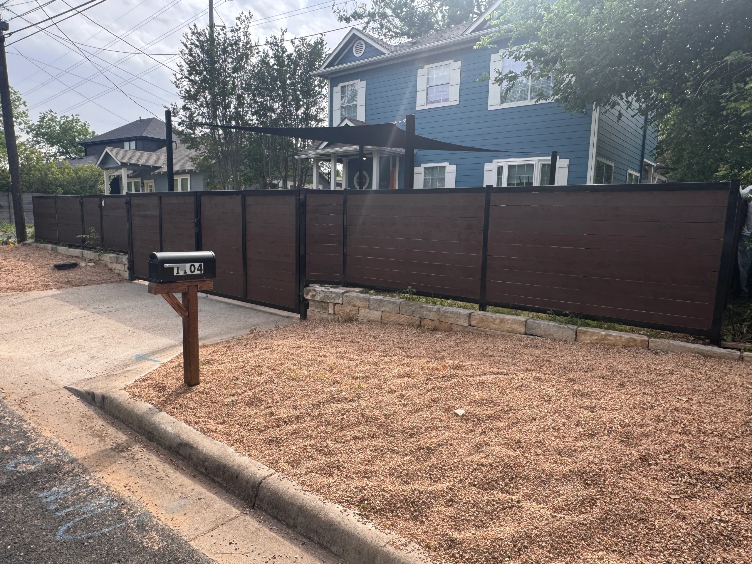 A black mailbox labeled 7104 stands near a gravel yard and sidewalk in front of a tall, dark wood fence—professionally finished by a local fence staining contractor—and a blue two-story house.