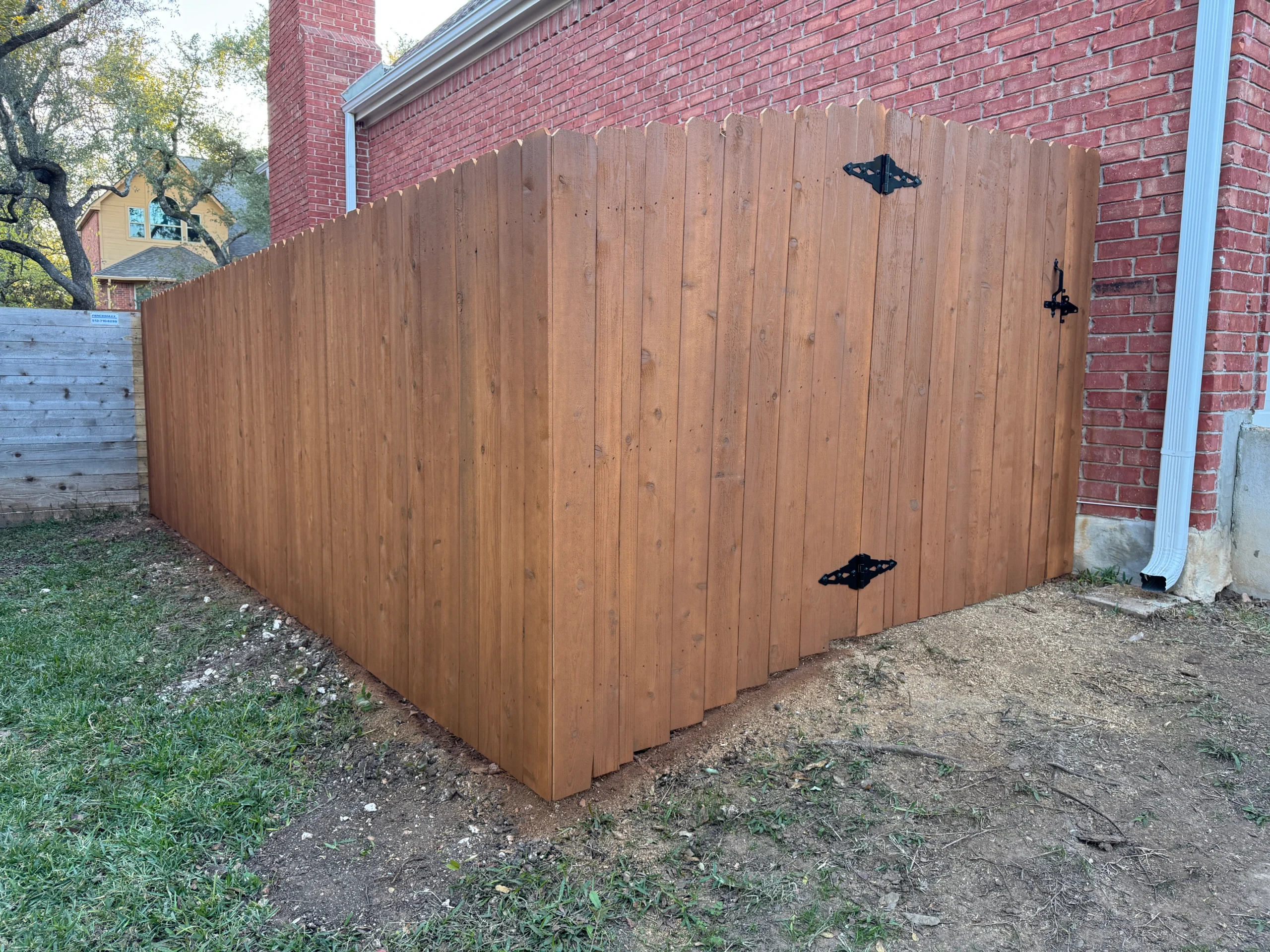 A wooden privacy fence with a gate and black metal hinges, expertly installed by a local fence staining contractor, runs alongside a red brick house. Grass and bare soil are visible in the foreground.