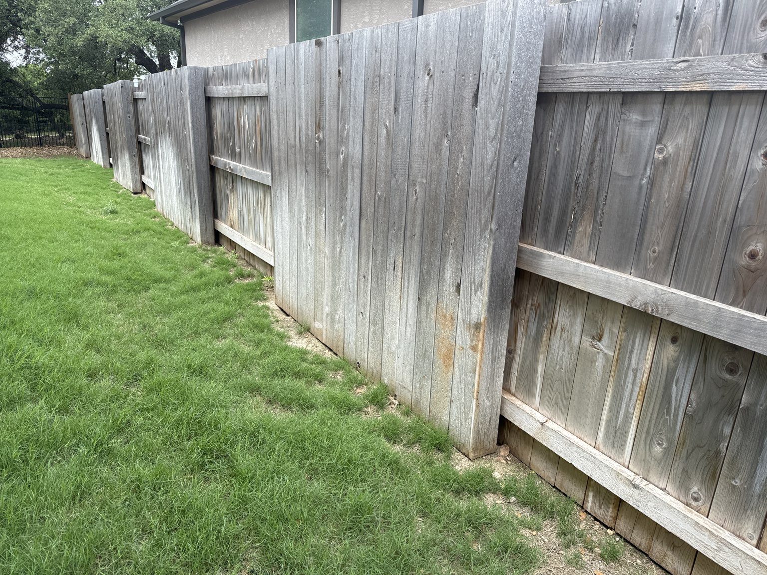A wooden privacy fence with leaning and uneven panels separates a grassy yard from a house.