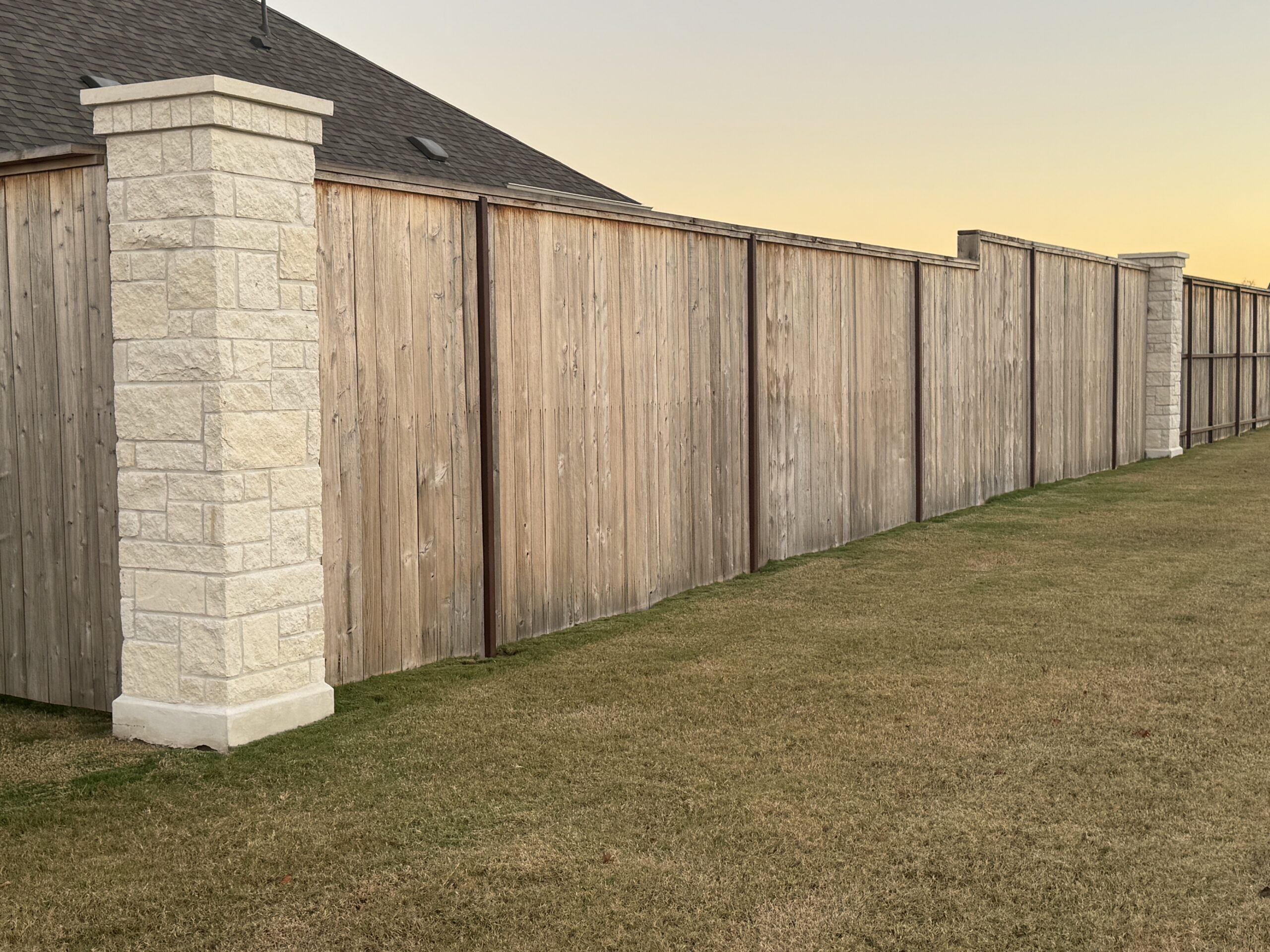 A wooden privacy fence with stone pillars separates a grassy yard from a neighboring house at sunset.