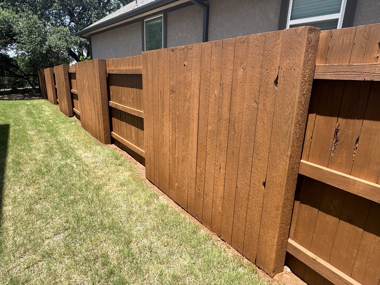 Wooden privacy fence with vertical planks runs alongside a house, dividing a grassy yard from neighboring properties under bright sunlight.