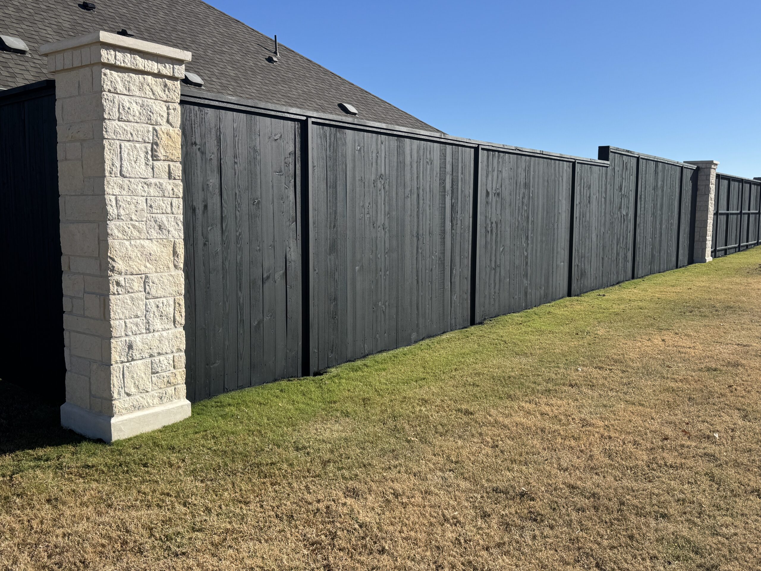 A dark wooden privacy fence with stone pillars stands along the edge of a backyard with dry, patchy grass under a clear blue sky.
