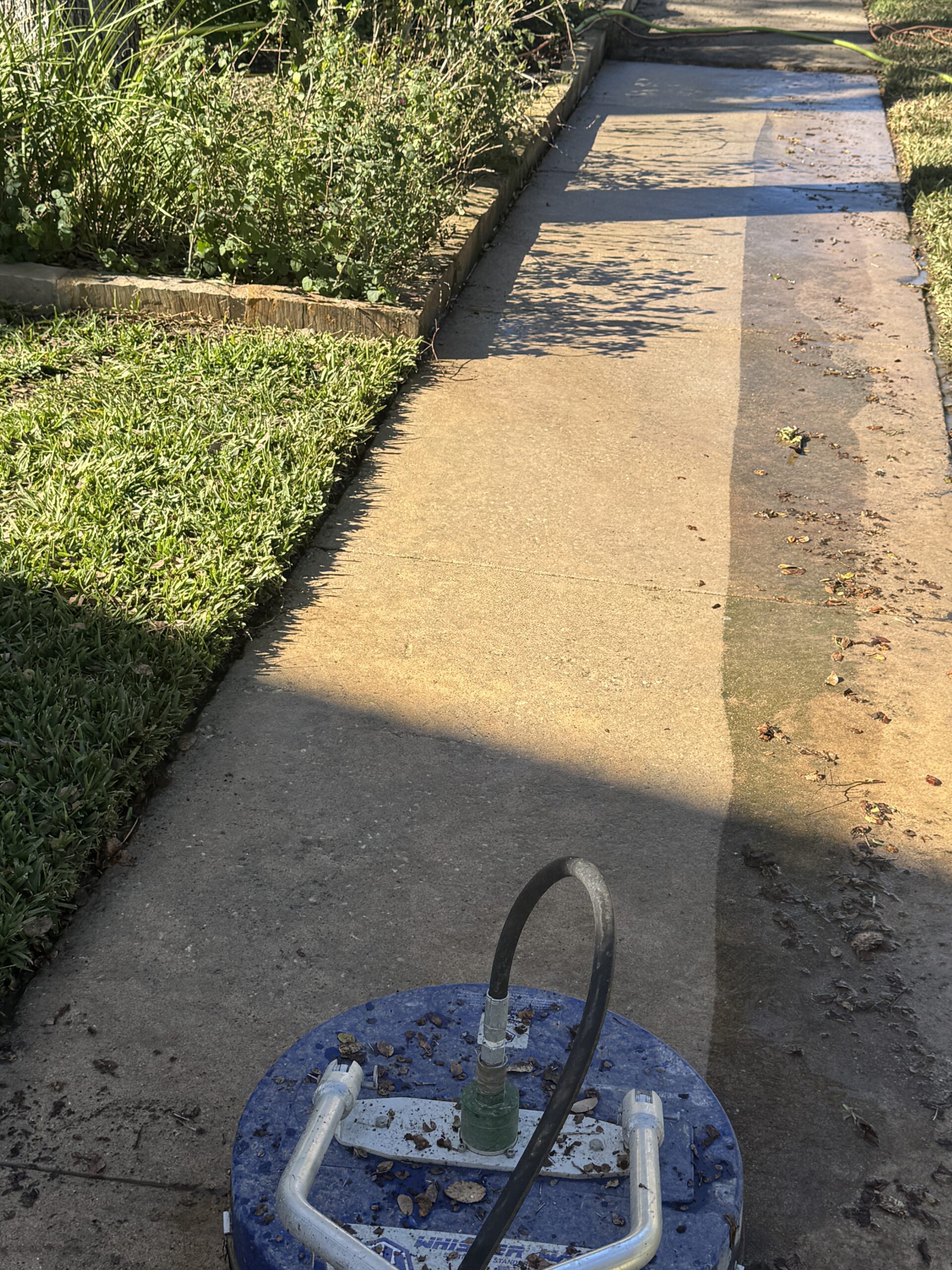 IMG_4289 A blue surface cleaner attached to a pressure washer is being used to clean a dirty concrete patio outdoors.