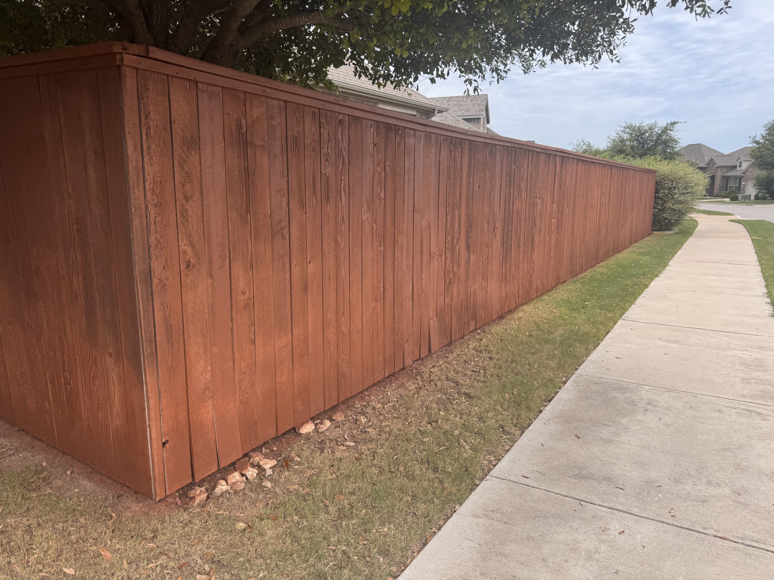 A long, reddish-brown wooden fence runs beside a sidewalk, bordered by grass and trees in a suburban neighborhood.