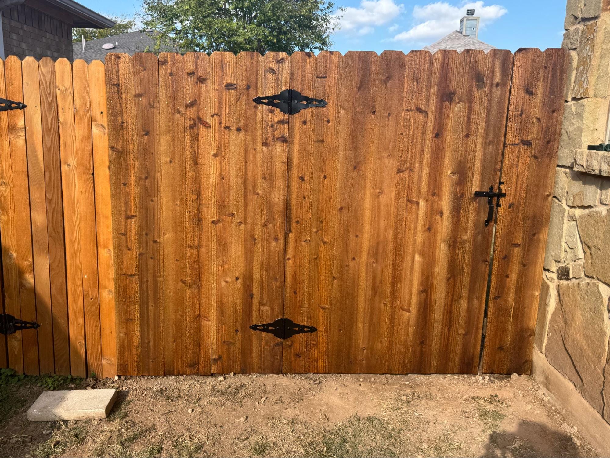 A closed wooden gate with black metal hinges and latch, set between a stone wall and a wooden fence, with a patch of bare ground in front.