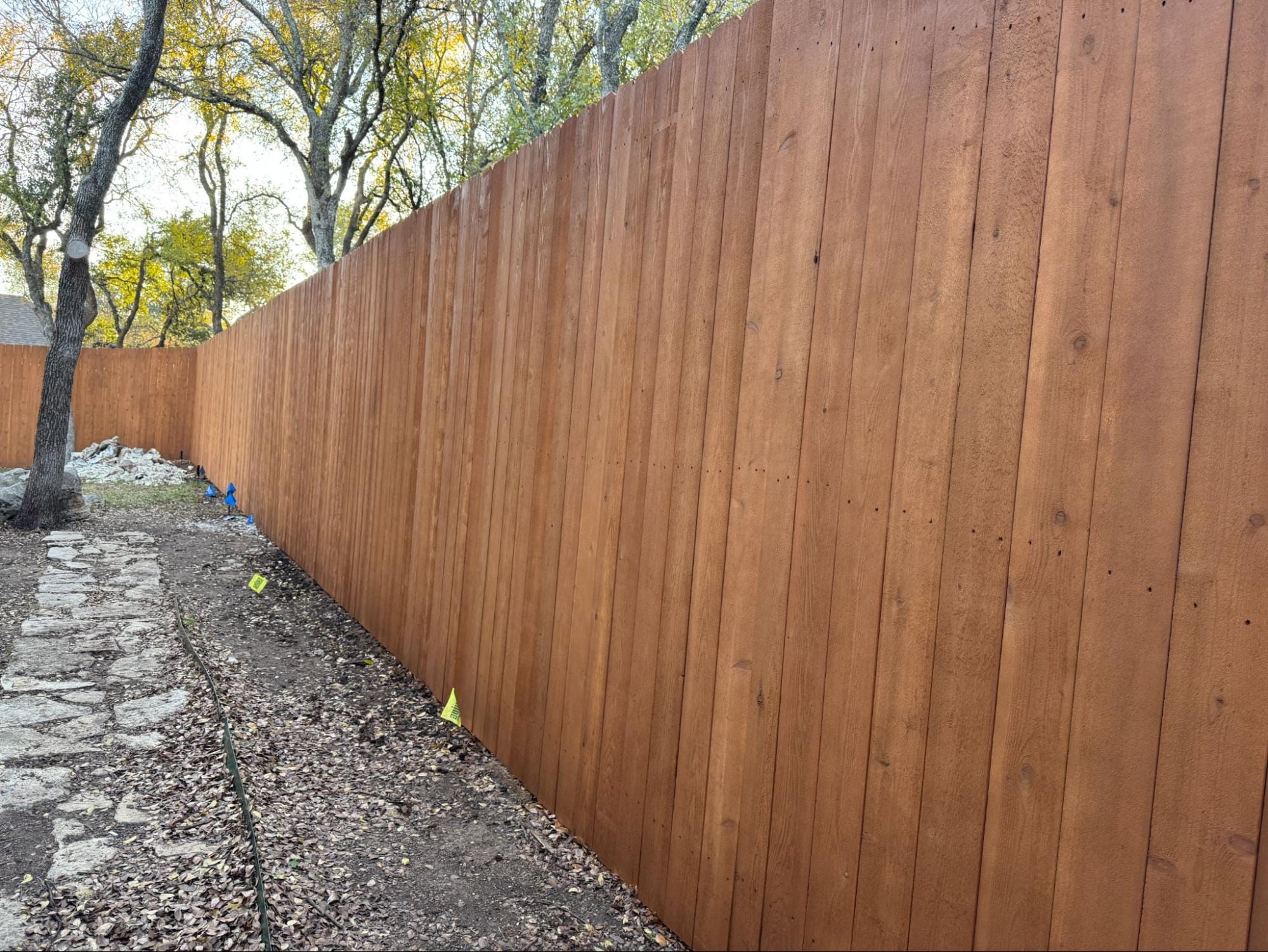 A tall wooden privacy fence runs alongside a dirt path with scattered leaves, bordered by trees with yellowing foliage.