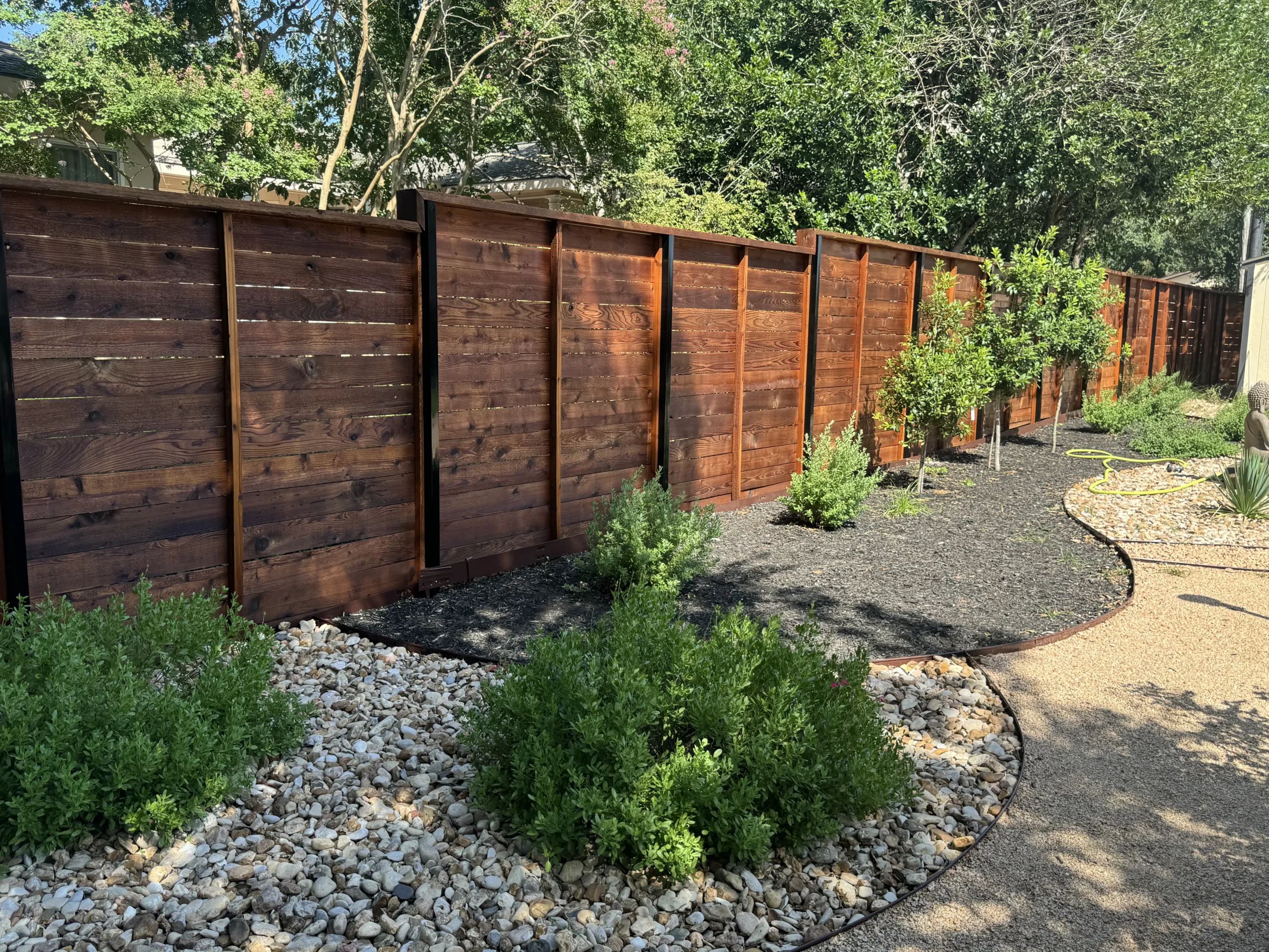 A wooden privacy fence, expertly maintained by a professional fence staining contractor, borders a landscaped yard with small trees, mulch, green shrubs, and a curved path with gravel and stone ground cover. Dense trees are visible behind the fence.
