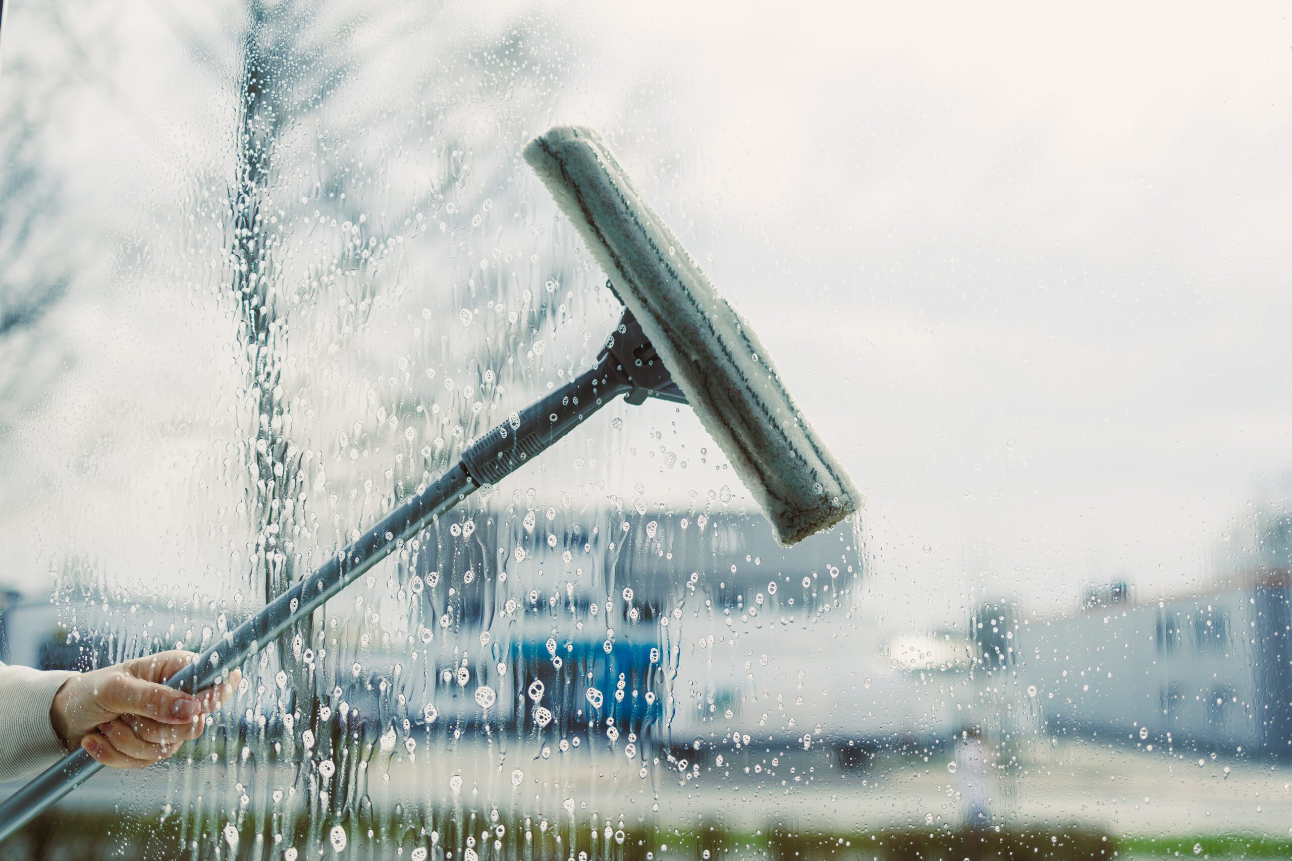 Glass cleaning with special chemicals. Cleaning service in a high-rise building. A person uses a squeegee to clean a large window covered in soap and water, with an outdoor scene blurred in the background.