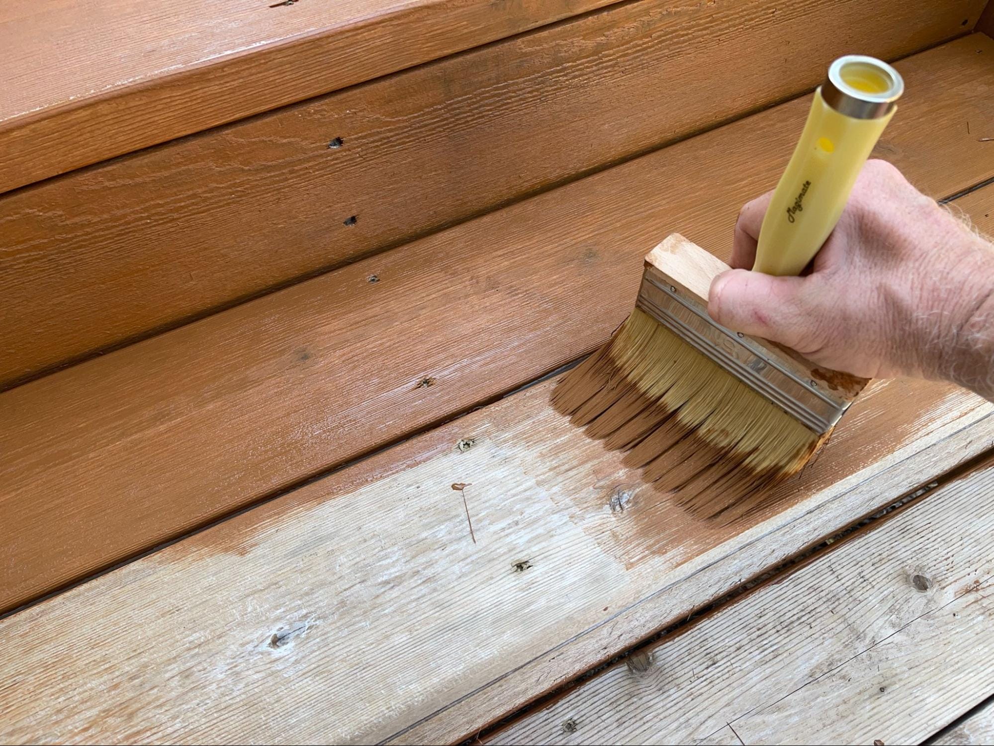 A hand uses a wide brush to apply wood stain or finish to outdoor wooden steps, covering a weathered area next to already stained wood.