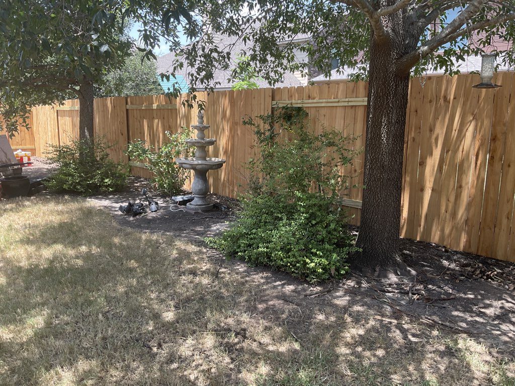 A wooden fence lines a backyard with trees, bushes, and a stone fountain. The grass is patchy and mostly dry.