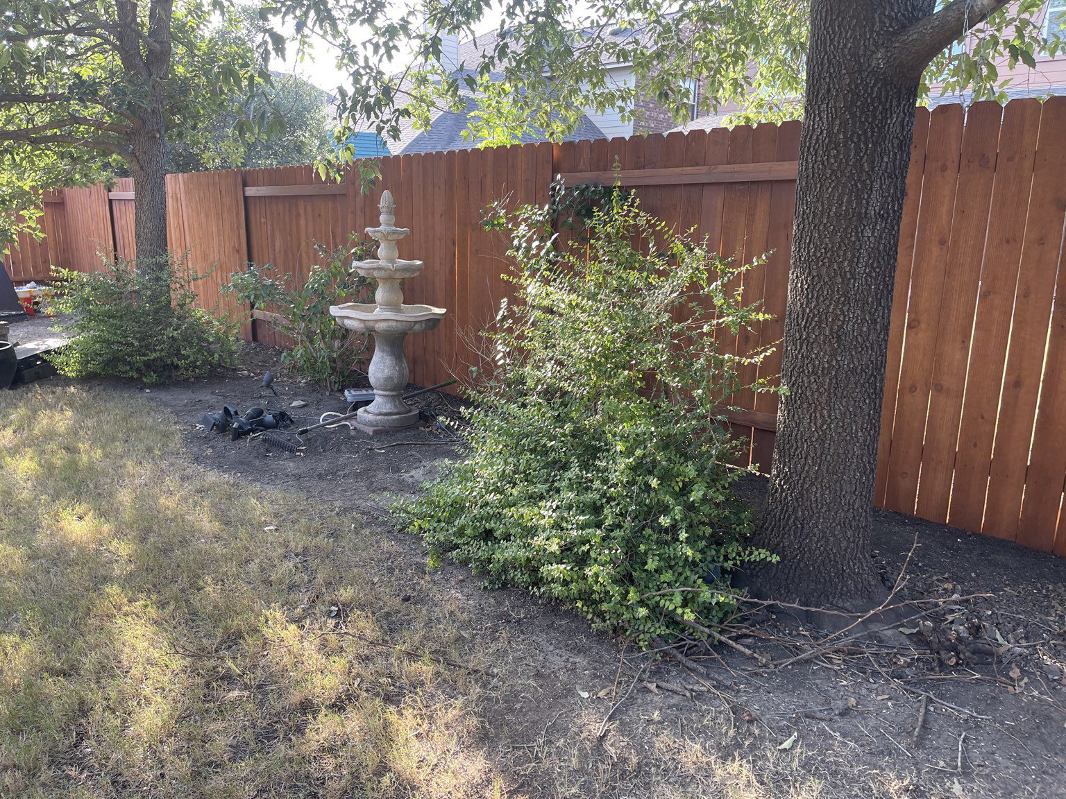 A stone fountain stands beside a leafy bush and tree in a backyard bordered by a tall wooden fence.
